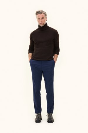 Ciemnobrązowy sweter z golfem, granatowe spodnie i czarne sznurowane buty z fakturowanymi sznurówkami. Model stoi z rękami w kieszeniach na neutralnym tle.