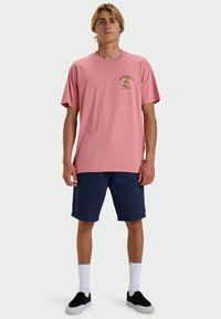 Jonge man staat in een roze T-shirt met een kleine afbeelding en marineblauwe shorts, witte sokken en zwarte instap schoenen.