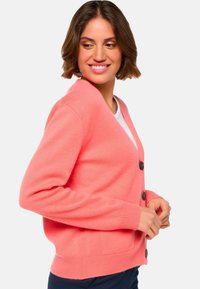 Femme souriante regardant sur le côté, portant un cardigan boutonné rose corail par-dessus un haut blanc avec un pantalon bleu marine, sur un fond uni.