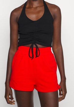 Person iført en ærmeløs sort crop top med frontbinding og lyse røde elastiske shorts med høj talje, stående mod en ensfarvet baggrund.