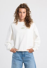 White sweatshirt with a high neckline and ribbed cuffs. Features a green embroidered logo on the chest. Paired with light blue jeans.