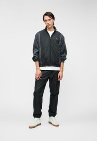 Μαύρο αθλητικό κοστούμι με ζακέτα με φερμουάρ και ταιριαστά παντελόνια. Διαθέτει λευκές λεπτομέρειες, ελαστικούς μανσέτες και ένα μικρό πράσινο λογότυπο στο στήθος.