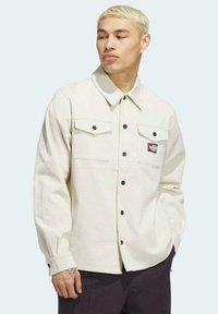 Camisa de pana de color beige claro con dos bolsillos en el pecho, botones negros y un pequeño parche con logo en el bolsillo izquierdo. Mangas largas y cuello clásico.