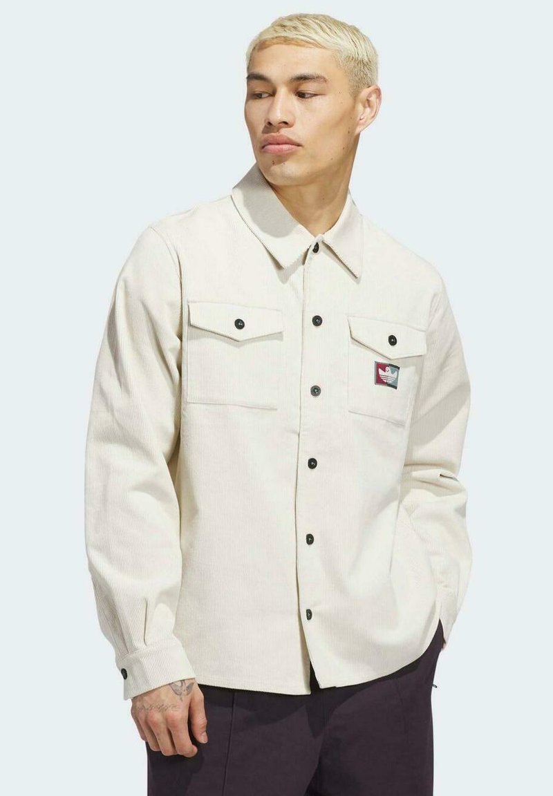 Camisa de pana de color beige claro con dos bolsillos en el pecho, botones negros y un pequeño parche con logo en el bolsillo izquierdo. Mangas largas y cuello clásico.