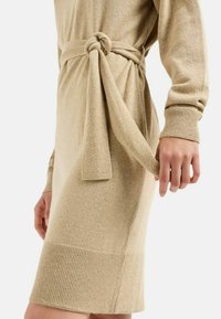 Abito lavorato a maglia color beige con vita cintata, maniche lunghe e orlo a coste. Il tessuto ha una consistenza morbida e una lucentezza sottile.