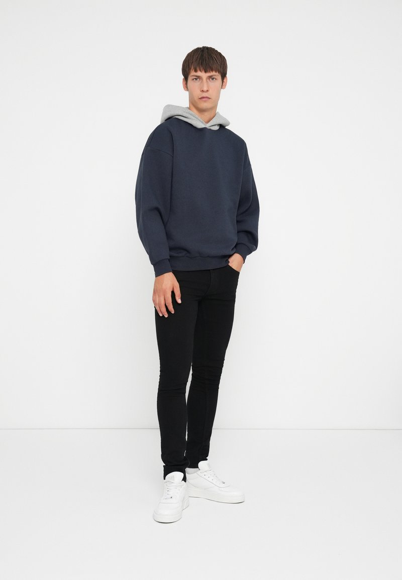 Navy Sweatshirt mit grauem Kapuzenaufsatz, lockerer Schnitt und fallenden Schultern; kombiniert mit schwarzen Skinny Jeans und weißen Turnschuhen.