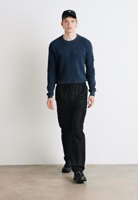 Maglione a coste blu navy, pantaloni cargo neri e scarpe nere. Modello che indossa un berretto nero, mentre cammina su uno sfondo chiaro e neutro.