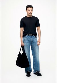 Mann står iført svart t-skjorte, blå jeans, svarte sko, og holder en stor svart handlenettpose mot en ensfarget hvit bakgrunn.