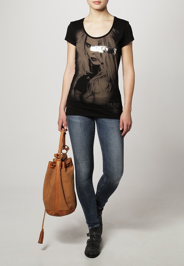 T-shirt noir à manches courtes avec imprimé graphique et détail en sequins, associé à un jean bleu et un sac seau en daim marron. Bottines noires.