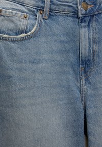 Gros plan sur un jean en denim bleu clair montrant la poche avant, la passants de ceinture, les coutures et un bouton métallique sur la ceinture.
