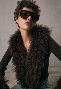 Gilet in finto pelliccia marrone, shaggy, indossato sopra un top aderente a maniche lunghe con costine. Modella che indossa occhiali da sole oversize con lenti gialle.