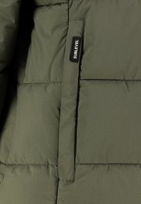 Nahaufnahme einer olivgrünen gepolsterten Jacke mit einer vertikalen Tasche und einem schwarzen rechteckigen Etikett mit der Aufschrift "SUBLEVEL."