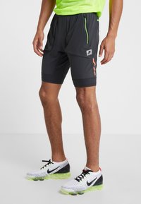 Man i svarta träningsshorts och vita Nike löparskor med neongröna sulor, stående på ett grått golv.