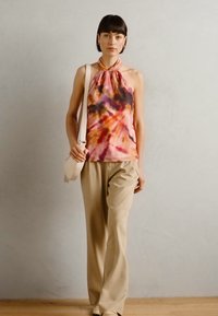Ärmelloses, mehrfarbiges Batik-Oberteil in rosa, orange und lila, kombiniert mit beigen, hochgeschnittenen Hosen. Lässige Umhängetasche.