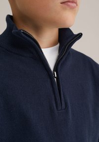 Mörkblå zip-up polotröja med hög krage, tillverkad av mjukt stickat material. Har en kontrasterande dragkedja och bärs över en vit T-shirt.
