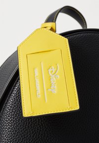 Juodas tekstūruotas kuprinės krepšys su geltona bagažo etikete, ant kurios iškilūs Disney ir Karl Lagerfeld logotipai, kontrastingai siuvinėtos detalės.
