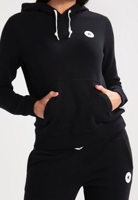 Sweat à capuche noir avec poche avant, cordon de serrage ajustable et logo étoile blanche. Confectionné en tissu doux avec poignets et ourlet côtelés.