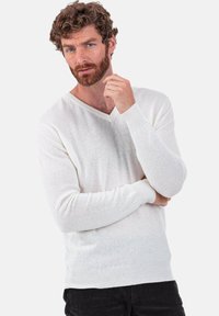 Weißer Strickpullover mit V-Ausschnitt, strukturierter Stoff sowie gerippte Bündchen und Saum. Das Modell steht mit der Hand am Kinn und schaut leicht von der Kamera weg.