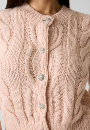 Zachtroze kabelgebreid vest met zilverkleurige sierknopen, voorzien van ingewikkelde gevlochten patronen en een ronde halslijn, gedragen door een persoon.