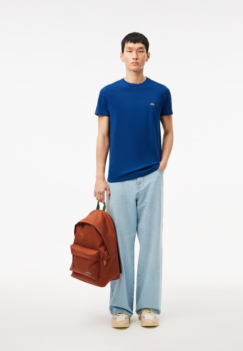 Blaues T-Shirt mit kurzen Ärmeln, lockere Passform, kombiniert mit hellblauen, weiten Jeans. Brauner Rucksack mit einer Fronttasche und grünen Akzenten. Lässige Schuhe.
