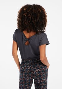 Dunkelgraues Oberteil mit kurzen Ärmeln und einem geknoteten Rückendetail. Kombiniert mit bunten, gemusterten Stretchbundhosen mit floralen Designs.