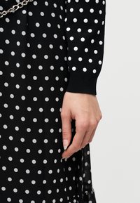 Χέρι που ακουμπά δίπλα σε μαύρο φόρεμα και πουλόβερ με λευκές πουά, με μια ασημένια αλυσίδα να διακρίνεται εν μέρει στο πάνω αριστερό μέρος.