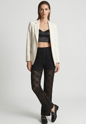 Kvinde stående iført en hvid blazer, sort satin cropped top, sorte bukser med blondemønster og sorte loafers mod en ensfarvet baggrund.