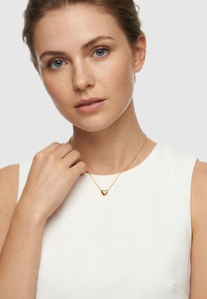 Frau mit heller Haut und blauen Augen, die ein weißes ärmelloses Oberteil und eine goldene herzförmige Anhängerkette trägt, Hand in der Nähe des Schlüsselbeins.