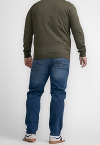 Man standing with back facing camera, wearing olive green sweater, blue jeans, and white sneakers with black stripes.