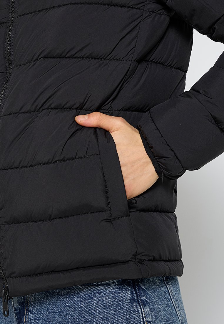 Chaqueta de plumas negra con textura acolchada, que cuenta con un bolsillo lateral. La tela parece suave y ligera, ideal para mantener el calor.