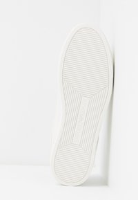 Vitt sneaker sula med horisontella spår och "Emporio Armani"-logotyp prägling i mitten, placerad vertikalt mot en vit bakgrund.