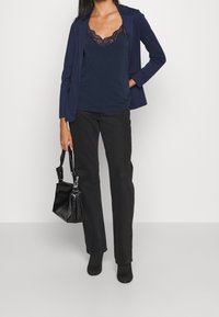 Tmavě modrý top s krajkovou výšivkou pod tmavě modrým sakem, doplněný černými kalhotami se rovnými nohavicemi a texturovanou černou kabelkou. Černé kotníkové boty doplňují celý vzhled.