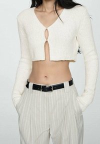 Pull côtelé crème cropped avec un col en V et une ouverture en forme de goutte à l'avant, associé à un pantalon rayé gris clair et une ceinture noire.