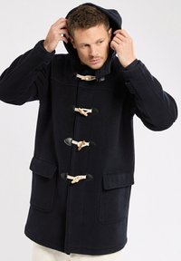 Homme ajustant la capuche d'un manteau noir en toile de jute avec des fermetures à bascule et des poches avant, sur un fond clair uni.