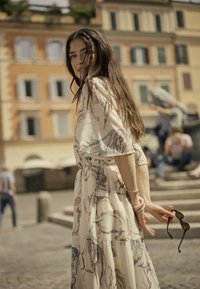 Világos bézs színű ruha virágmintákkal, V-nyakkal, karcsúsított derékkal és bő ujjakkal, napszemüveggel a kézben. A háttérben épületek láthatók.