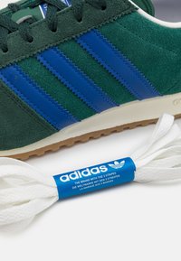 Zielony zamszowy sneaker z trzema niebieskimi paskami i białą podeszwą, wyposażony w białe sznurowadła owinięte niebieską taśmą z logo adidas.