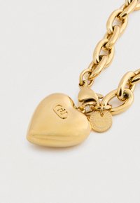 Χρυσό βραχιόλι αλυσίδας με μενταγιόν σε σχήμα καρδιάς και μικρή στρογγυλή ετικέτα με εγκεφαλισμένα αρχικά μάρκας, τοποθετημένο σε λευκό φόντο.