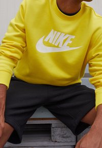 Gul sweatshirt med vitt Nike-logga på framsidan; tyget är mjukt och slätt. Kombinerad med svarta shorts, som visar en avslappnad stil.