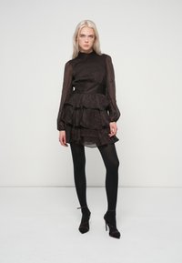 Μαύρο φόρεμα με στρώσεις, ψηλό λαιμό, μακριά διάφανη μανίκια και φούστα με βολάν. Το μοντέλο φοράει μαύρες κολάν και γυαλιστερά τακούνια.