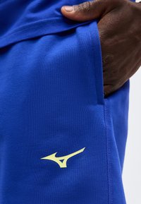 Σκούρο δέρμα χέρι που στηρίζεται μέσα στην τσέπη του φωτεινού μπλε αθλητικού σορτς με λογότυπο Mizuno σε ανοιχτό κίτρινο κοντά στη ραφή.