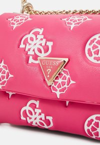 Sac à main rose Guess avec des motifs de logo blancs et une bandoulière chaîne dorée, avec une plaque métallique triangulaire du logo Guess U.S.A. sur le rabat avant.