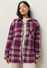 Camicia di flanella a quadri nei toni del viola, rosa e blu. Presenta una chiusura frontale con bottoni, colletto, maniche lunghe e una tasca sul petto. Tessuto morbido.