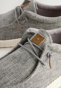 Chaussures décontractées en tissu gris tissé avec des lacets élastiques gris clair, des semelles blanches tachetées et un patch en cuir brun avec un embossage de soleil sur la languette.