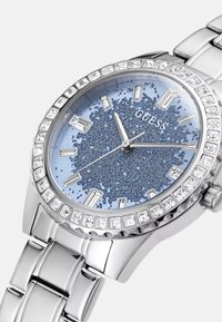 Srebrny zegarek ze stali nierdzewnej z okrągłą niebieską tarczą z brokatem, kryształowymi znacznikami godzin oraz diamentową obręczą. Posiada bransoletę z ogniwami.