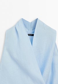 Chemise en lin bleu clair avec un large col en V et une étiquette de marque noire visible à l'intérieur du col.