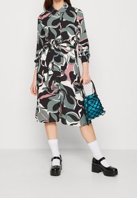 Vícebarevné šaty s listovým vzorem, dlouhými rukávy a stahovacími šňůrkami v pase, doplněné lesklou tyrkysovou kabelkou a botami na platformě.