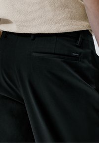 Mustat housut, jotka on valmistettu sileästä kankaasta. Housuissa on sivutasku ja vyötärön yksityiskohdat. Materiaali on pehmeäpintainen ja siinä on hienovarainen kiilto.