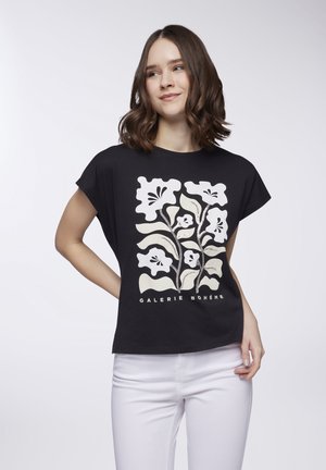 T-shirt nero a maniche corte con stampa floreale bianca e testo "GALERIE BOHÈME" nella parte inferiore. Abbinato a pantaloni bianchi.
