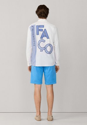 Hombre con camisa blanca de manga larga con un gran texto azul "FASCONNABLE" y pantalones cortos azules, de pie frente a un fondo liso de color gris claro.
