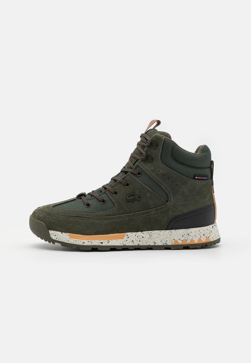 Chaussure de randonnée montante en vert foncé avec fermeture à lacets sur le devant, col de cheville rembourré, semelle blanche et orange tachetée, et petit logo sur le côté.